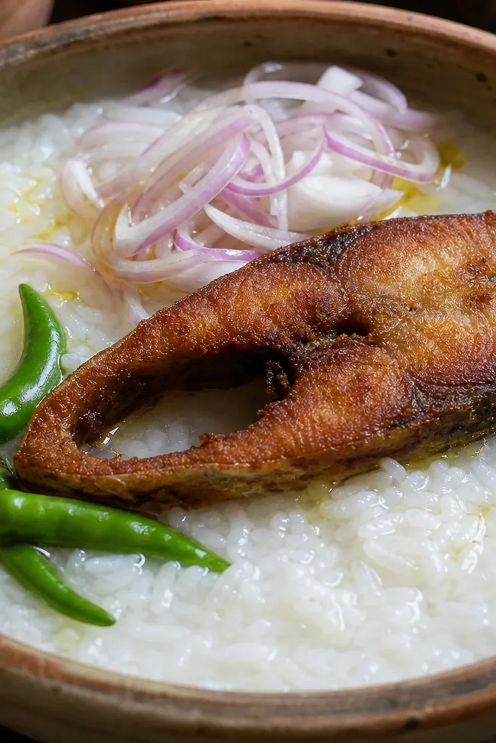 Bowl of Panta Bhat with fried Hilsa fish, garnished with green chilies and onions, showcasing vibrant colors and textures.
