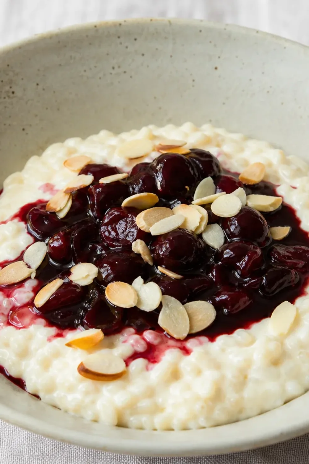 A bowl of creamy rice pudding topped with bright red cherry sauce and slivered almonds, garnished with mint.