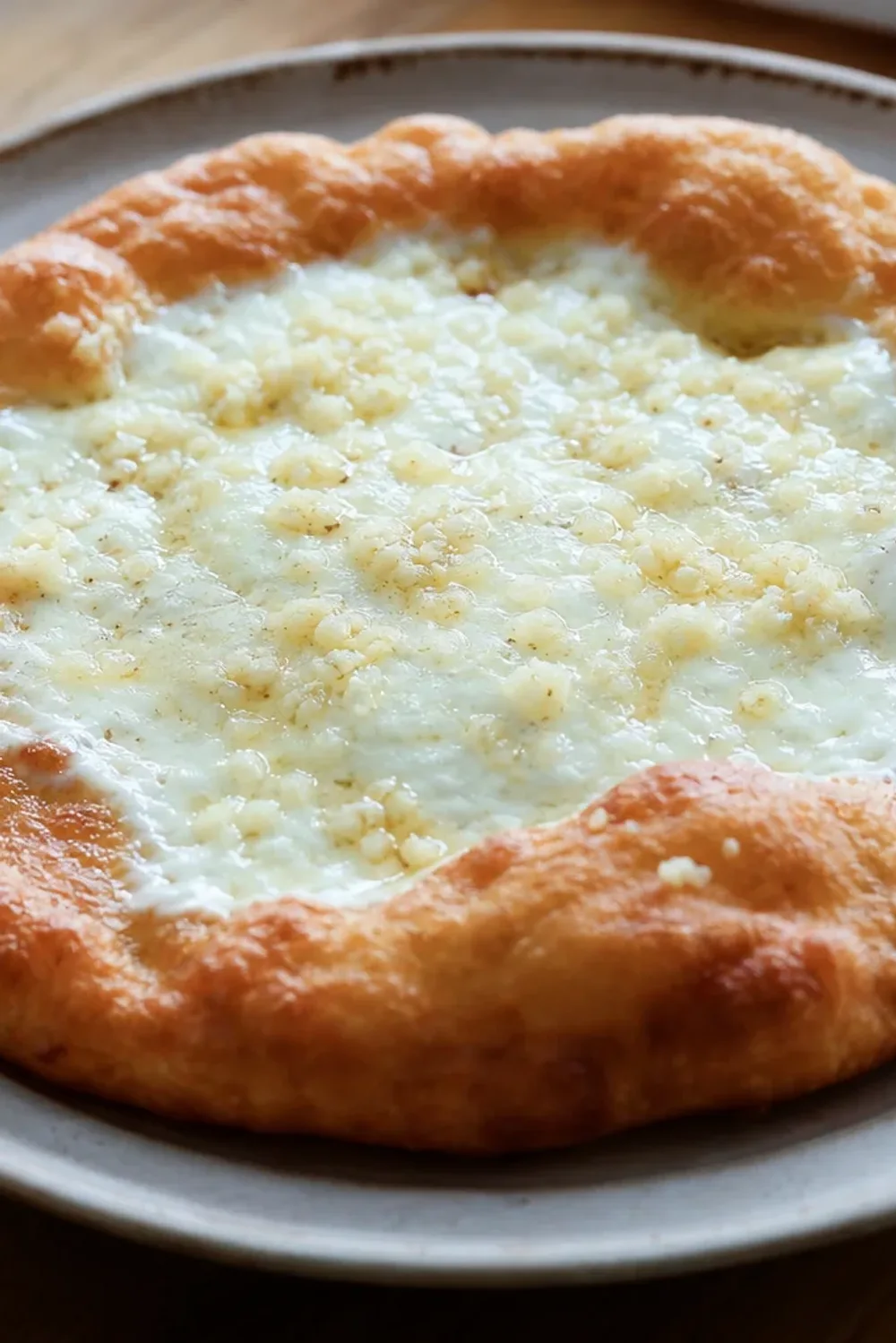 Golden-brown lángos topped with creamy sour cream, melted cheese, and garlic, garnished with herbs on a rustic plate.