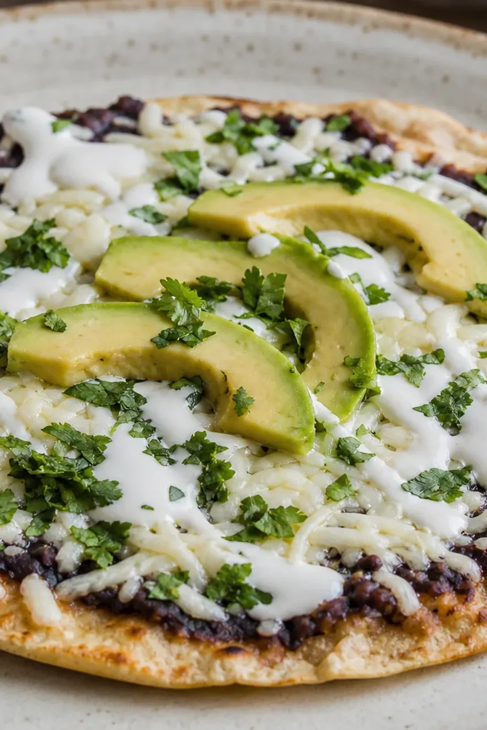 Crispy tlayuda topped with black beans, melted cheese, and sliced avocado, garnished with fresh cilantro.